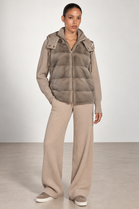 A model stands in a studio wearing a taupe-brown puffer vest over a matching beige zip-up hoodie and beige wide-leg knit pants, paired with beige sneakers.