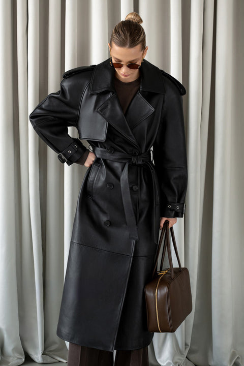 A woman wears a genuine black leather trench coat featuring a double-breasted silhouette, notched lapels, and belted cuffs crafted from soft buttery leather for a stylish and functional look.