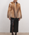 A model stands against a plain off-white studio wall wearing a voluminous tan and brown fur coat over a white top, paired with a black high-slit skirt and black pointed-toe heels.