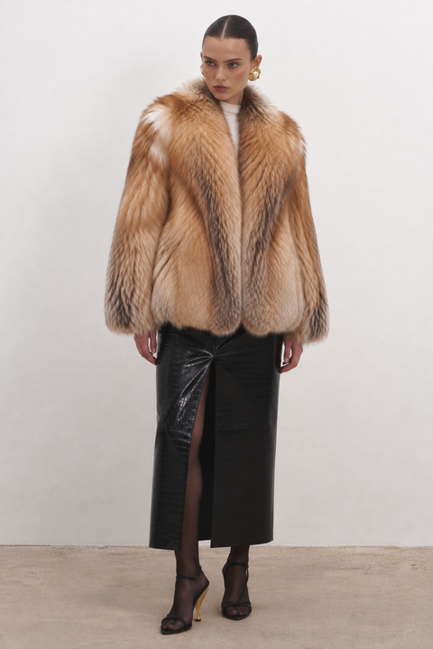A model stands against a plain off-white studio wall wearing a voluminous tan and brown fur coat over a white top, paired with a black high-slit skirt and black pointed-toe heels.